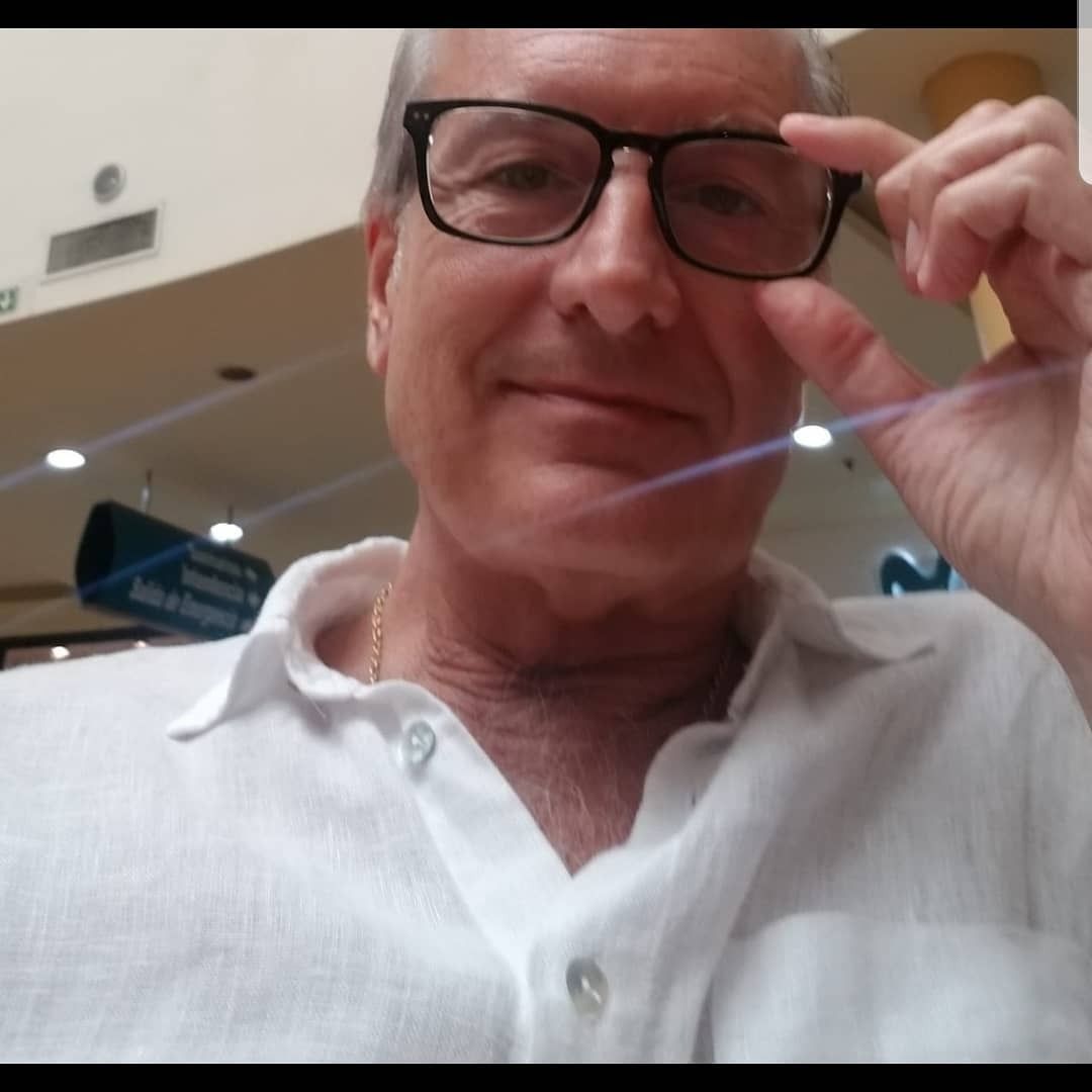 Una persona con camisa blanca se ajusta unas gafas negras en el interior de un edificio, con una leve sonrisa.