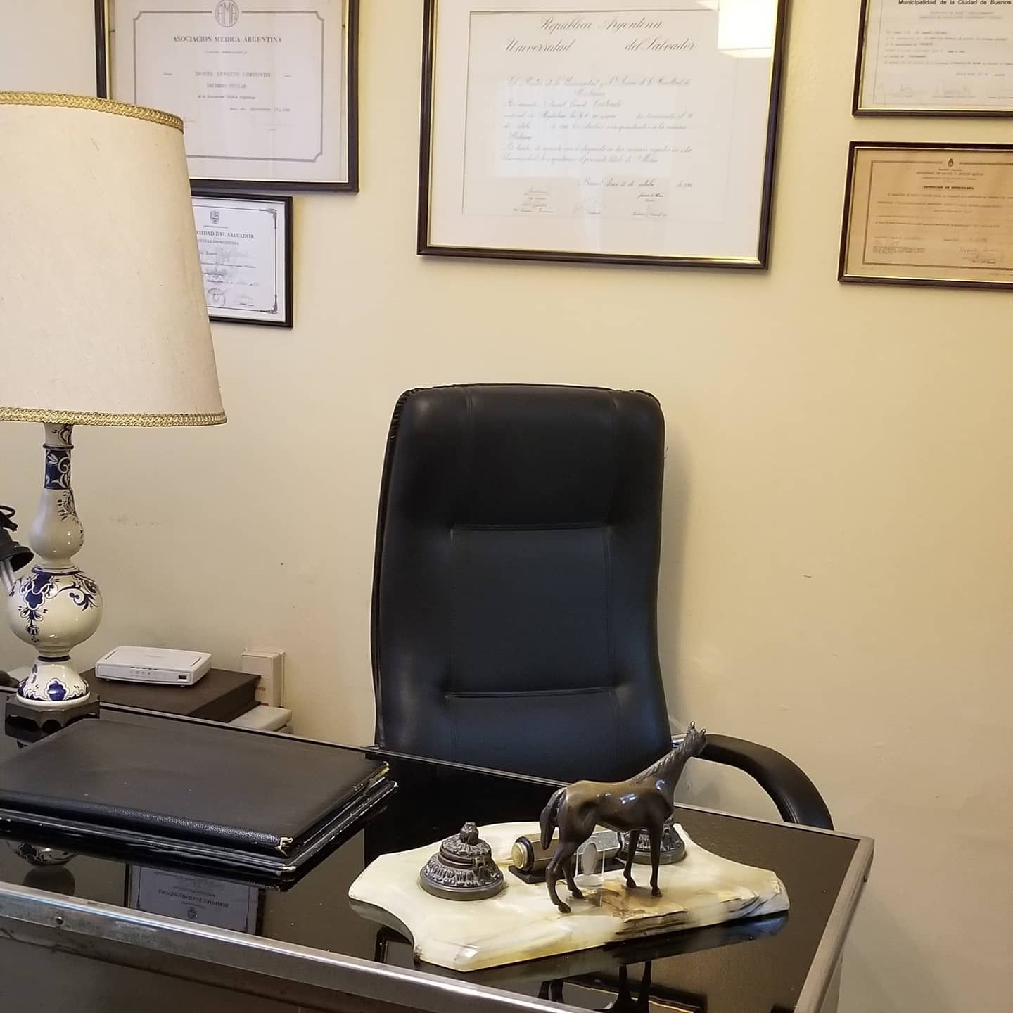 Oficina vacía con silla negra, certificados enmarcados, lámpara de escritorio y figuritas decorativas sobre una mesa.