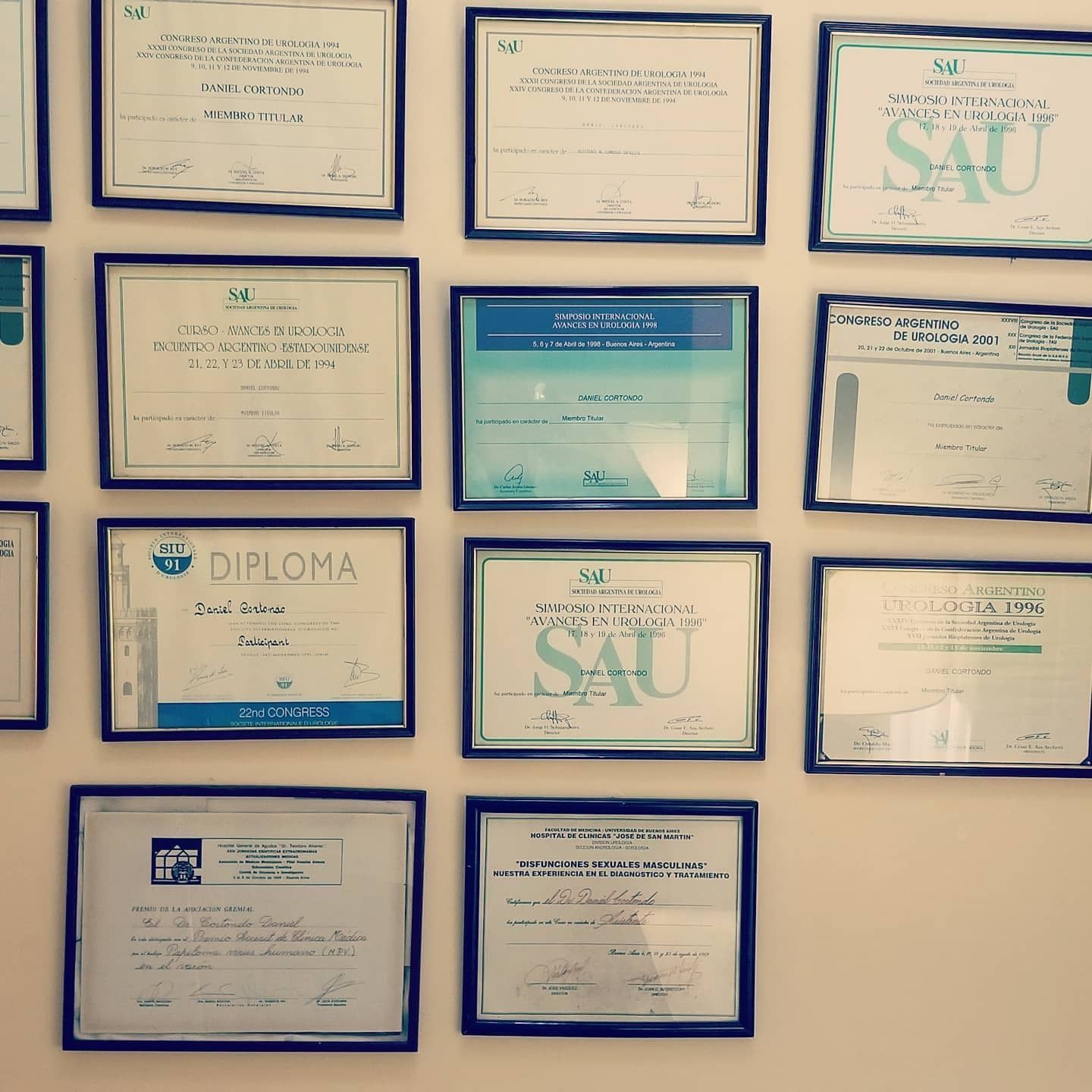 Pared de certificados y diplomas enmarcados en marcos azules, turquesas y beige sobre una pared beige.