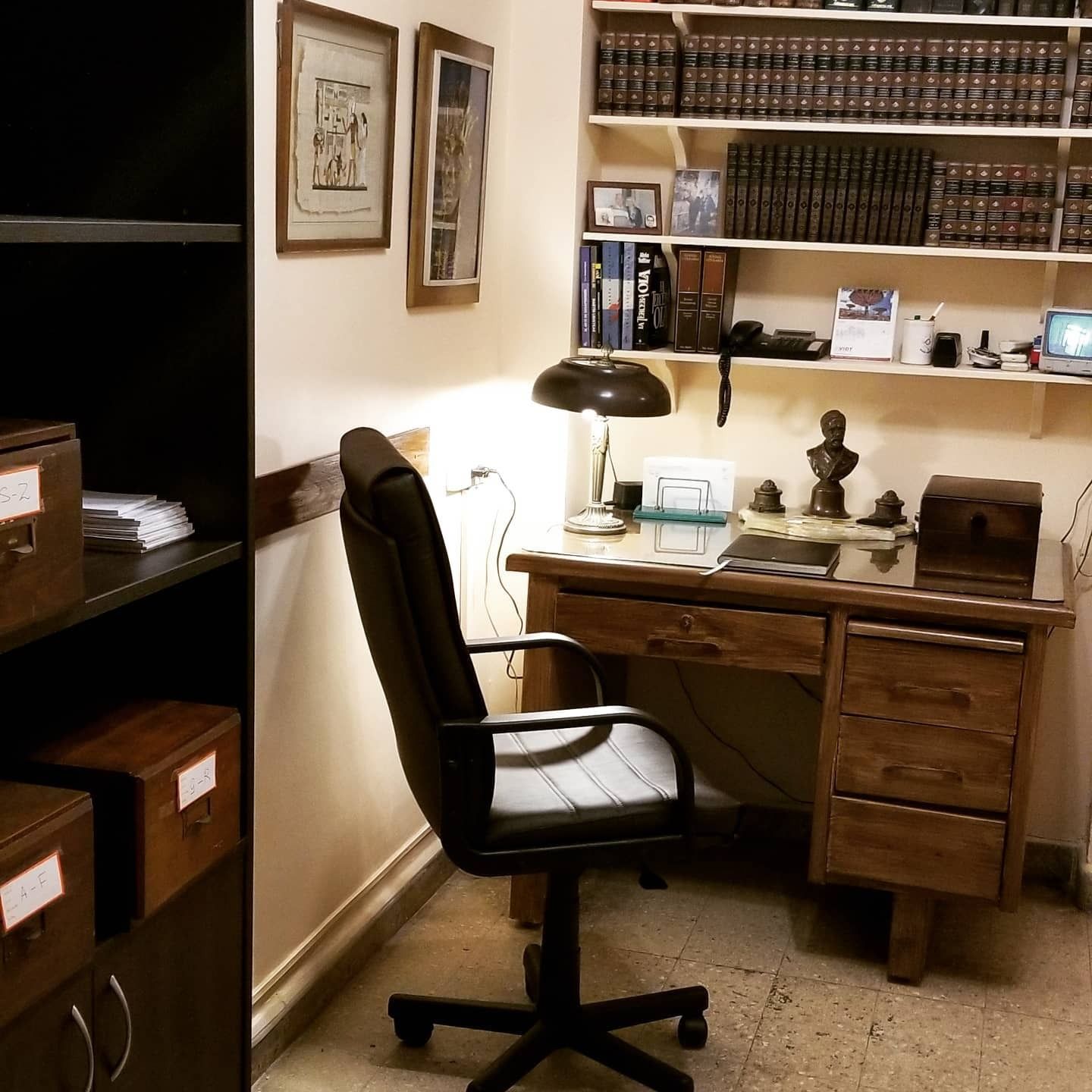 Oficina en casa con silla negra, escritorio de madera, lámpara de escritorio, estanterías y cuadros enmarcados.