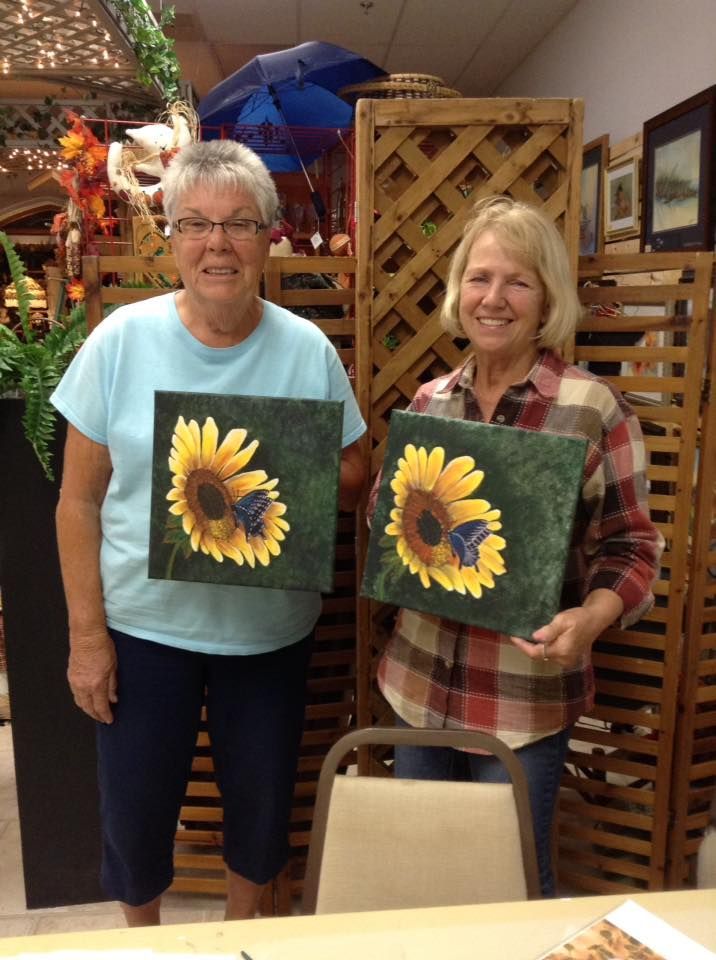 Two person standing next to each other holding paintings of sunflowers