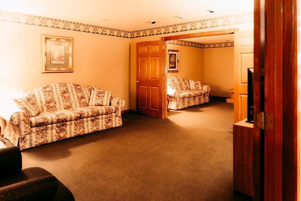 Warm beige living room with patterned sofas, carpet, framed art, and a doorway leading to another seating area