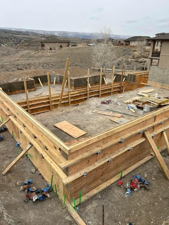 Wooden forms set for a concrete foundation on a hillside construction site, with a distant residential area.