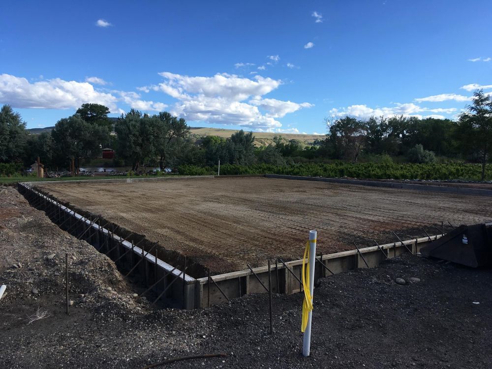 Concrete foundation with rebar, dirt, and gravel, under a blue sky with clouds.