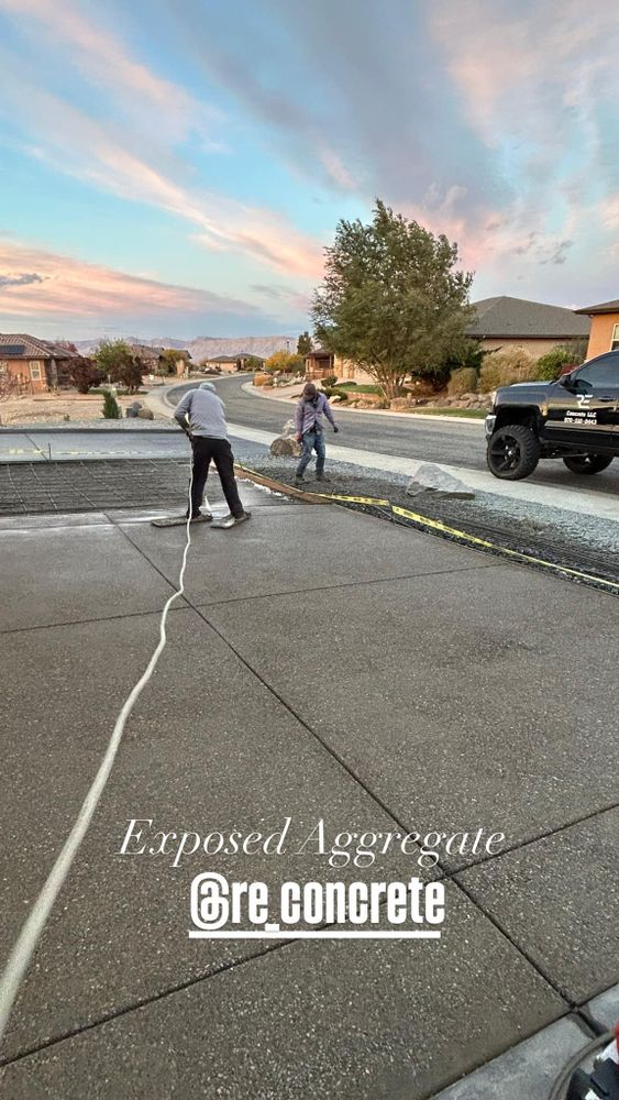 Two people cleaning exposed aggregate concrete on a street with a hose and brushes.