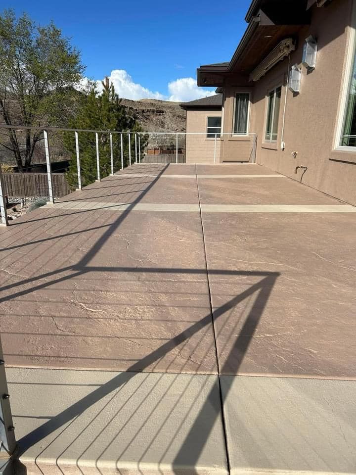 Exterior view of a deck with brown and beige concrete and a glass railing, with a house in the background.