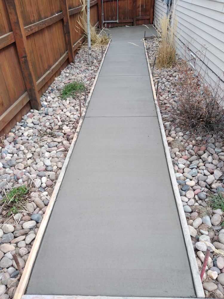 New concrete walkway between a wooden fence and a house, bordered by small rocks and wood.