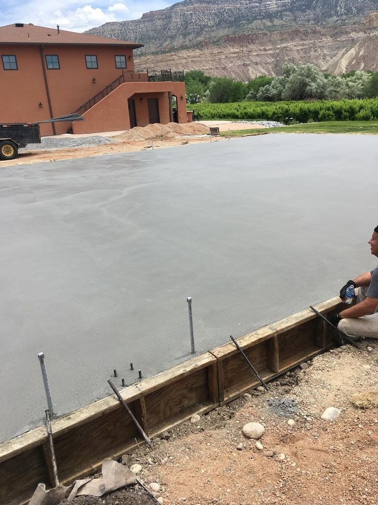 Freshly poured concrete surface with worker kneeling at edge, wooden formwork, building in background.