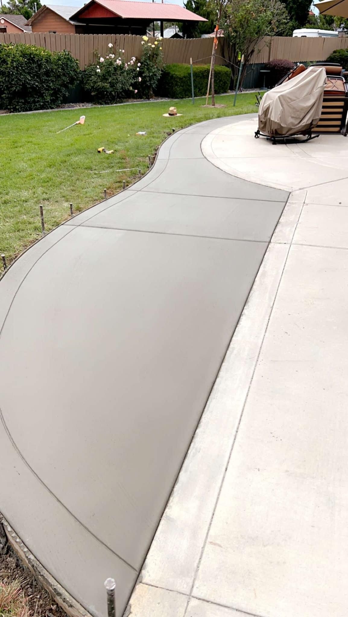Freshly poured concrete walkway curving through a grassy yard, next to an existing concrete patio.