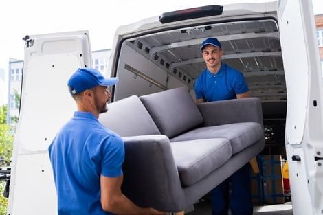 Two movers in blue uniforms loading a gray couch into a white van.