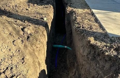 Trench dug in dirt, with green pipe visible. Sunlit.