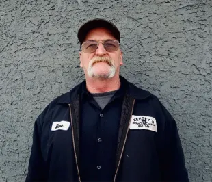Man with mustache wearing a cap and work jacket, standing in front of a gray wall.