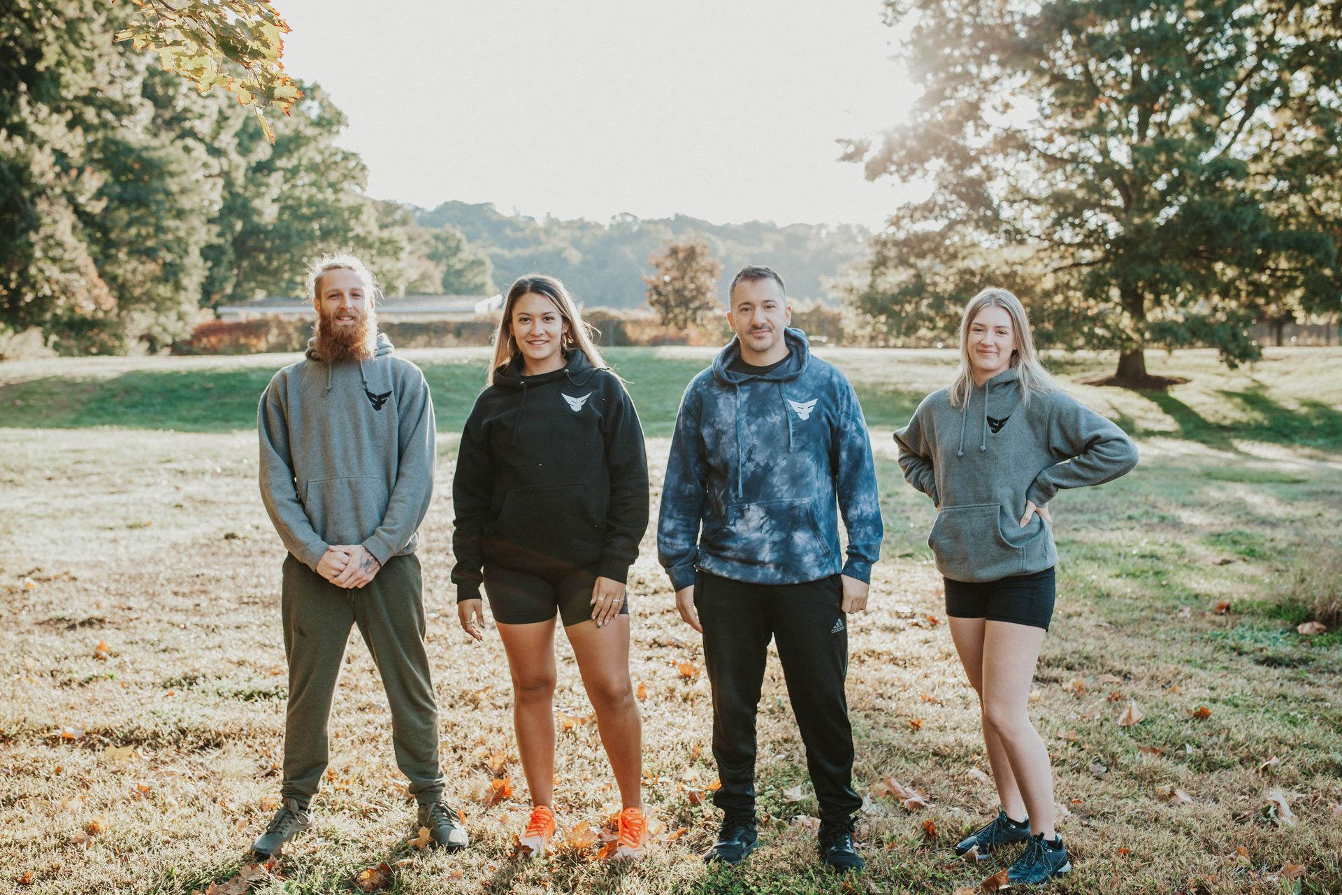 Four people wearing hoodies stand in a grassy field. Sunlight shines through trees.