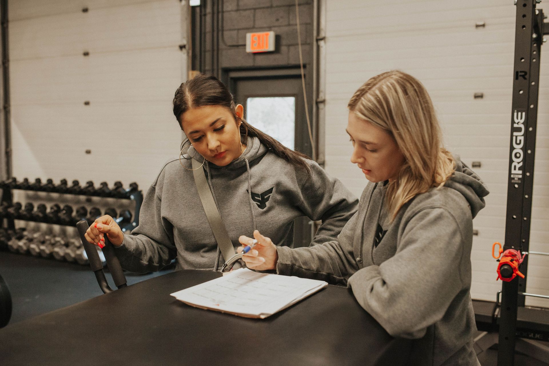 Two women in gray hoodies reviewing paperwork in a gym setting.