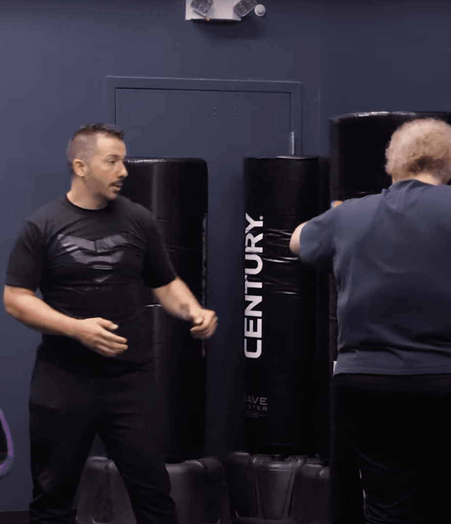 Man in black shirt demonstrates a punch while another person punches a Century punching bag in a gym.