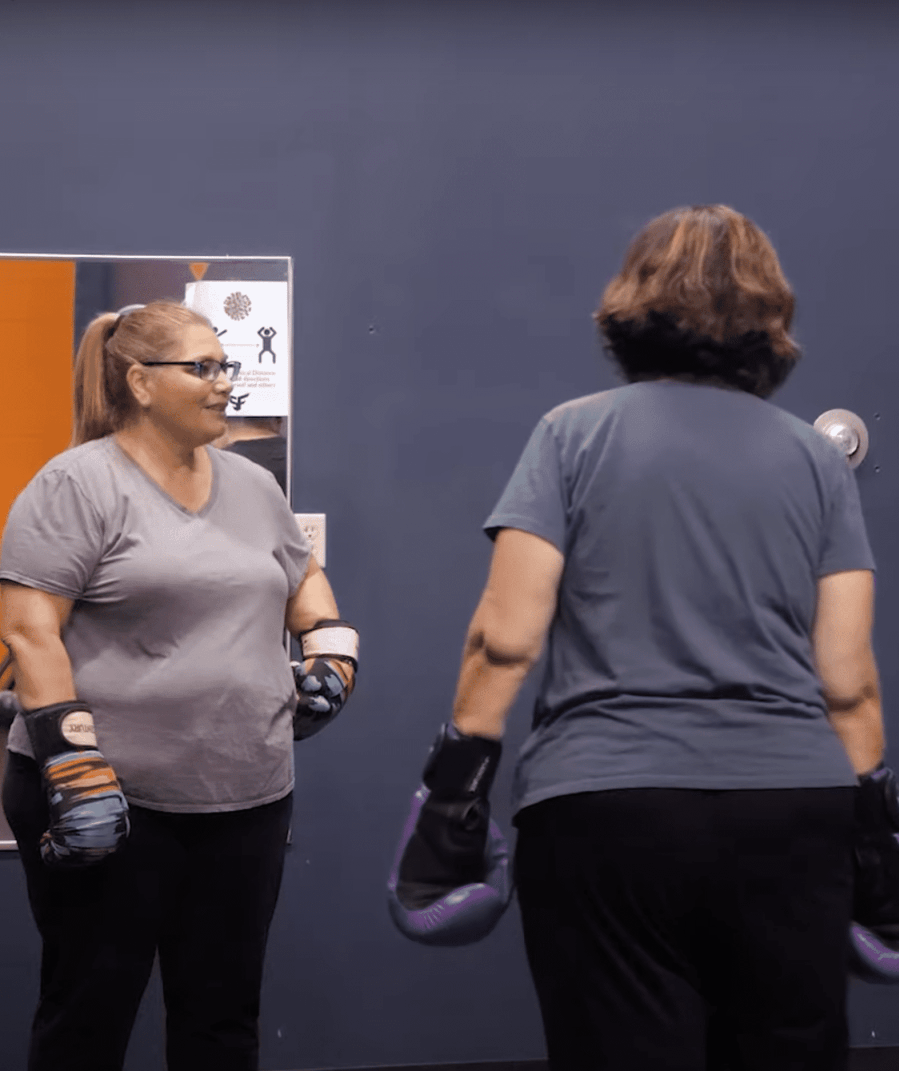 Two women in boxing gloves, one facing the camera, smiling, in a gym.