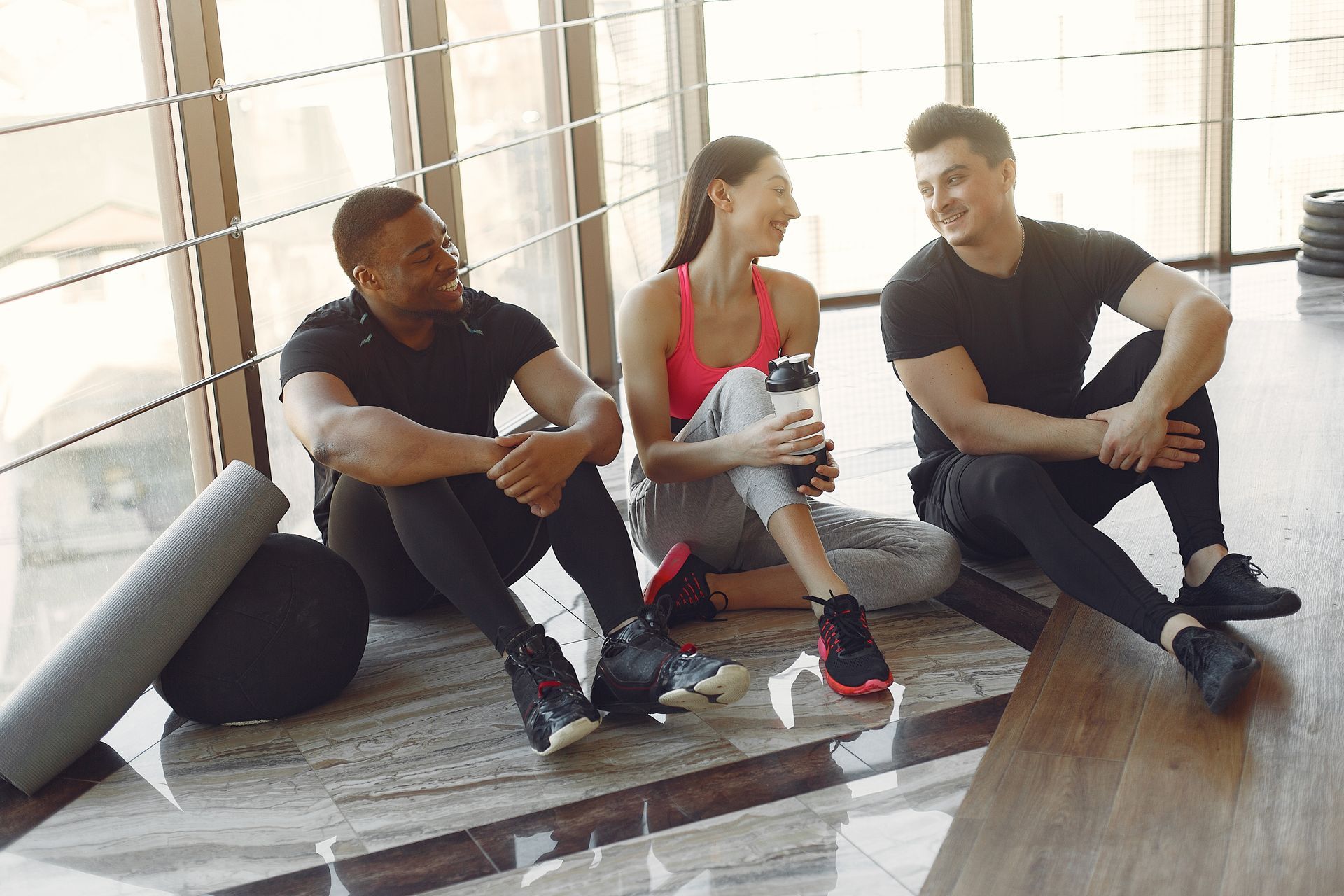 Three people in workout clothes sitting and chatting on a gym floor with large windows.