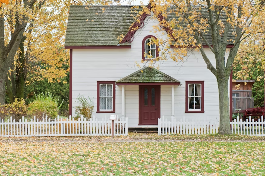 White cottage with red trim and picket fence amid autumn trees and fallen leaves