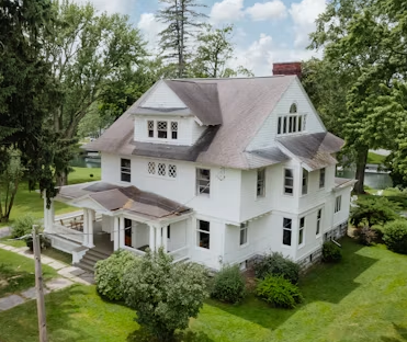 White farmhouse with a porch on a green lawn, surrounded by trees under a cloudy sky