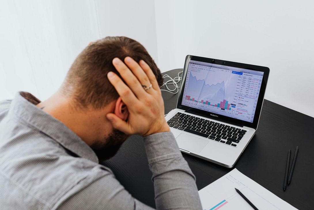 Person holding head at desk beside laptop showing a falling stock chart