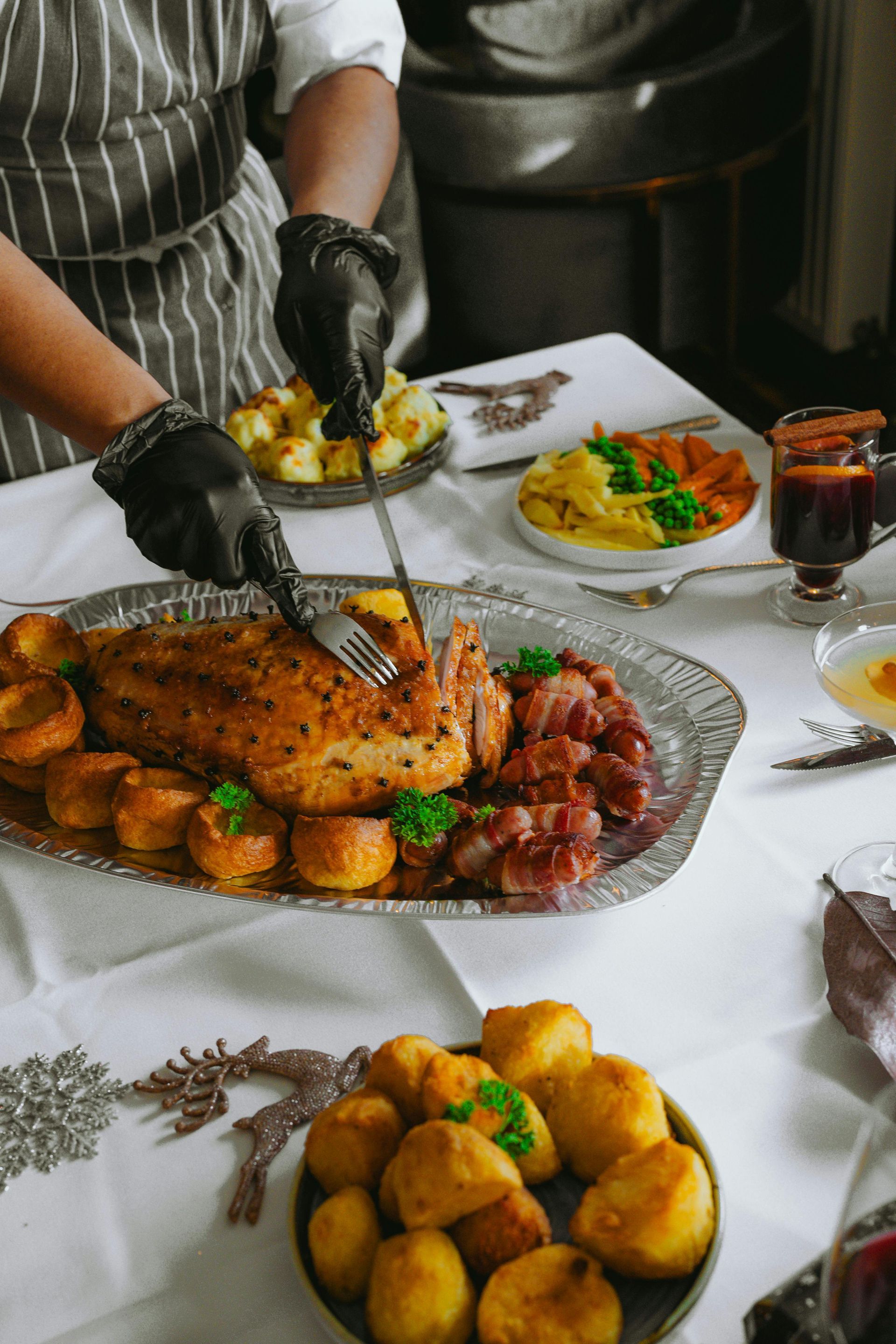 Person serving roasted meat and potatoes at a dining table with salads and drinks