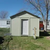 A beige, rectangular storage shed with a white door stands in a grassy yard under a clear blue sky.