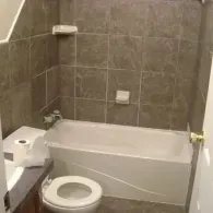 A bathroom featuring a white bathtub, a toilet in the foreground, and dark brown tiled walls with built-in shelves.