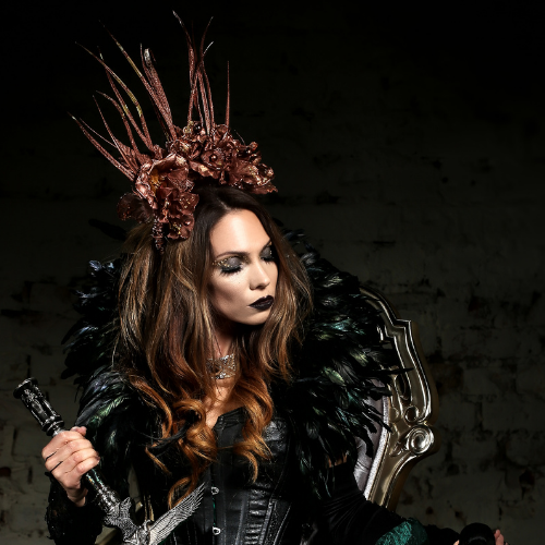 Woman in black feathered attire and ornate crown, holding a sword against a dark background.