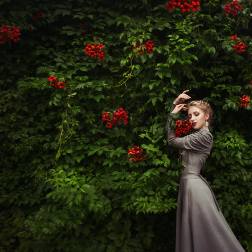 Woman in a gray dress posing beside a lush green wall with clusters of red flowers