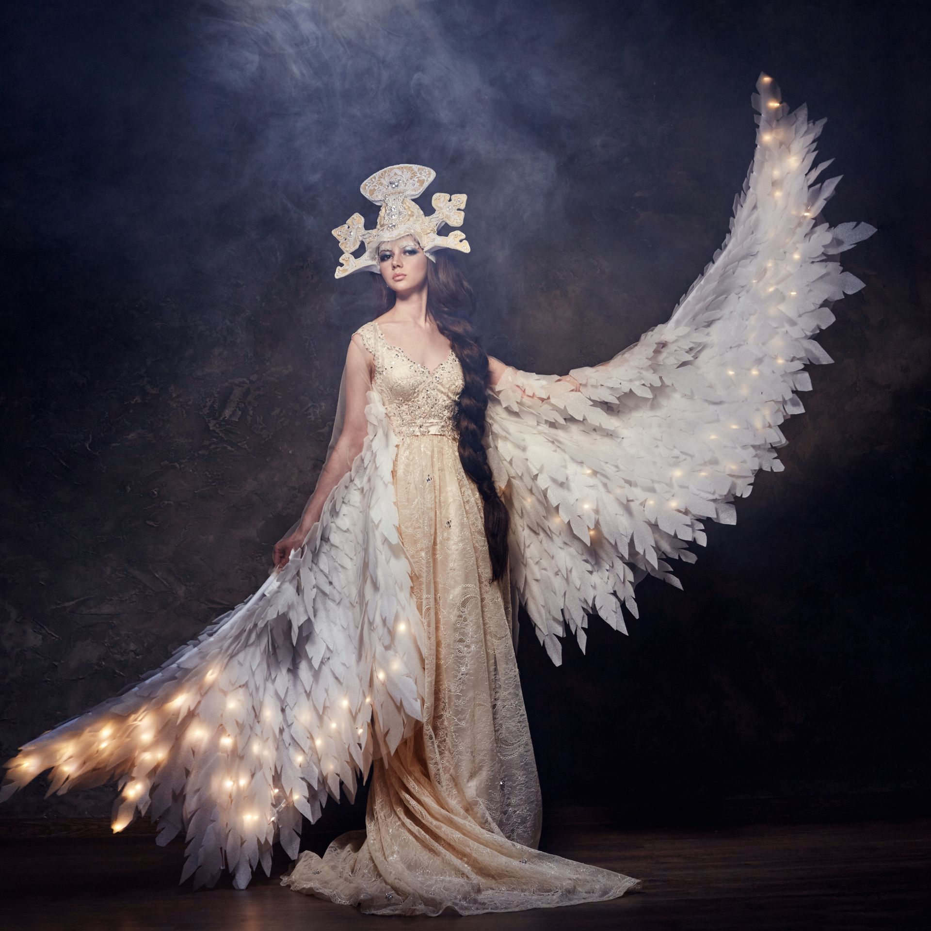 Woman in an ornate white gown with feathered wings and crown, standing against a dark smoky backdrop