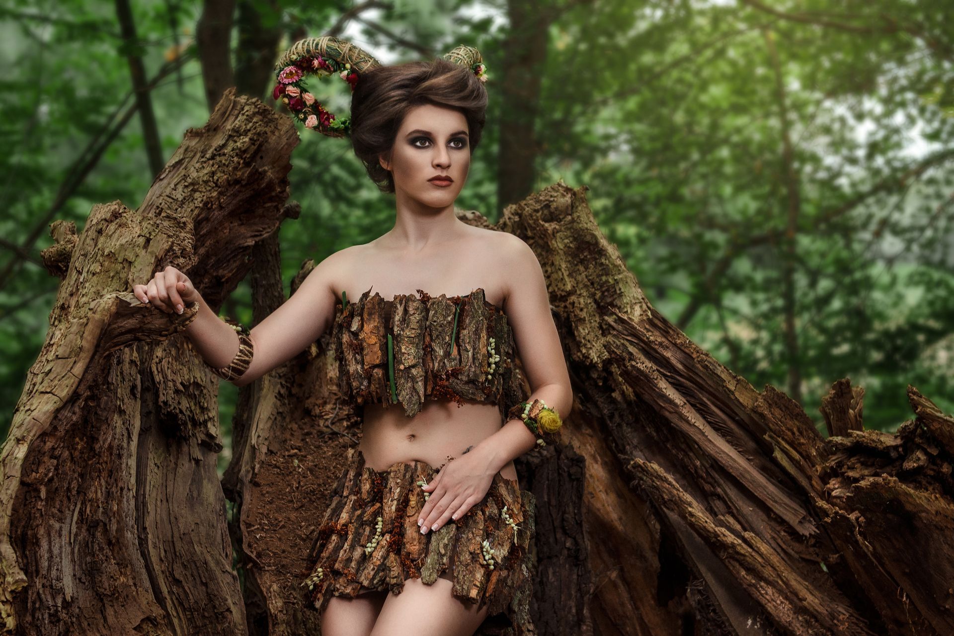 Woman in a leafy bark dress posing in a forest, framed by large tree roots.