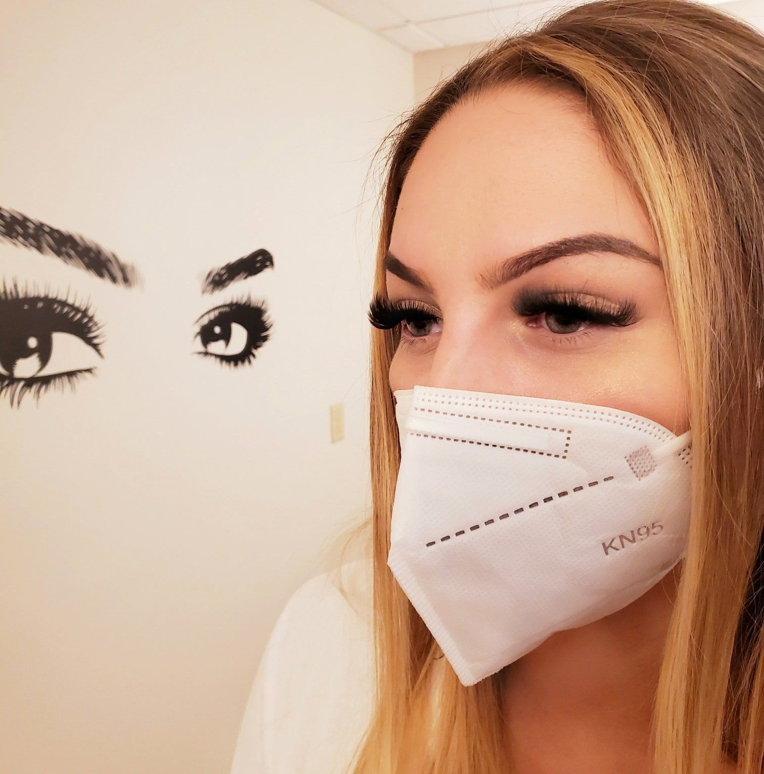 Woman with long lashes and mask next to wall art of eyes.
