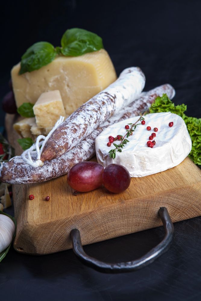 Tagliere di formaggi con salsicce a fette e intere, cracker, uva e un formaggio a pasta molle su un tagliere di legno.