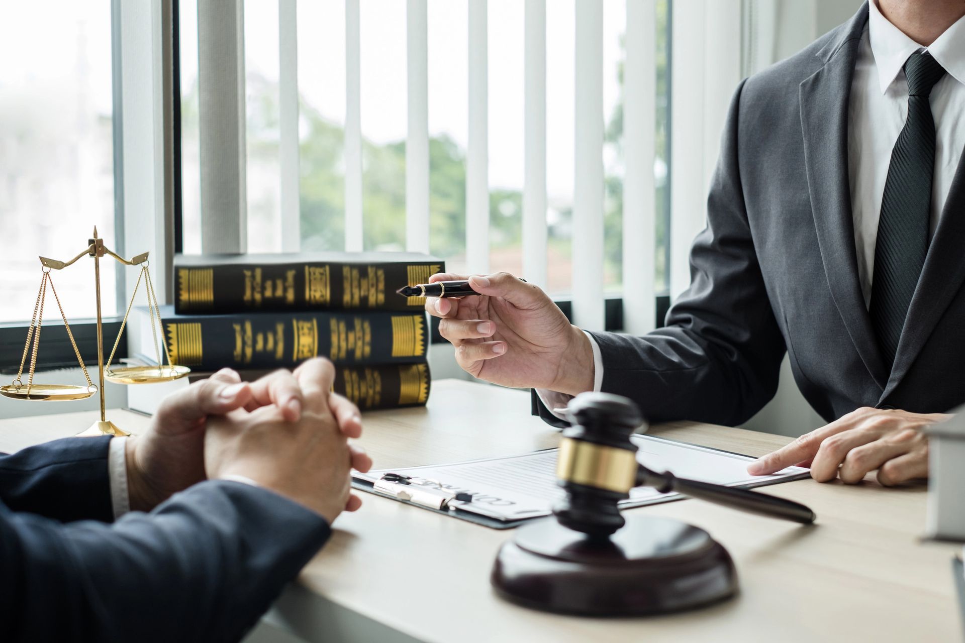 Un professionista legale in giacca e cravatta gesticola mentre si consulta con un cliente dall'altra parte di una scrivania, dove ha con sé un martelletto e dei libri di diritto.