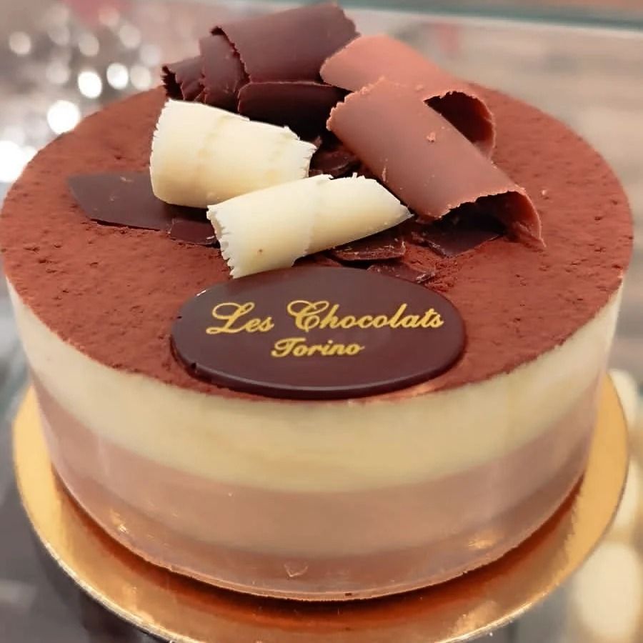 Torta al cioccolato a tre strati con cacao in polvere, scaglie di cioccolato e un logo su una targhetta dorata.