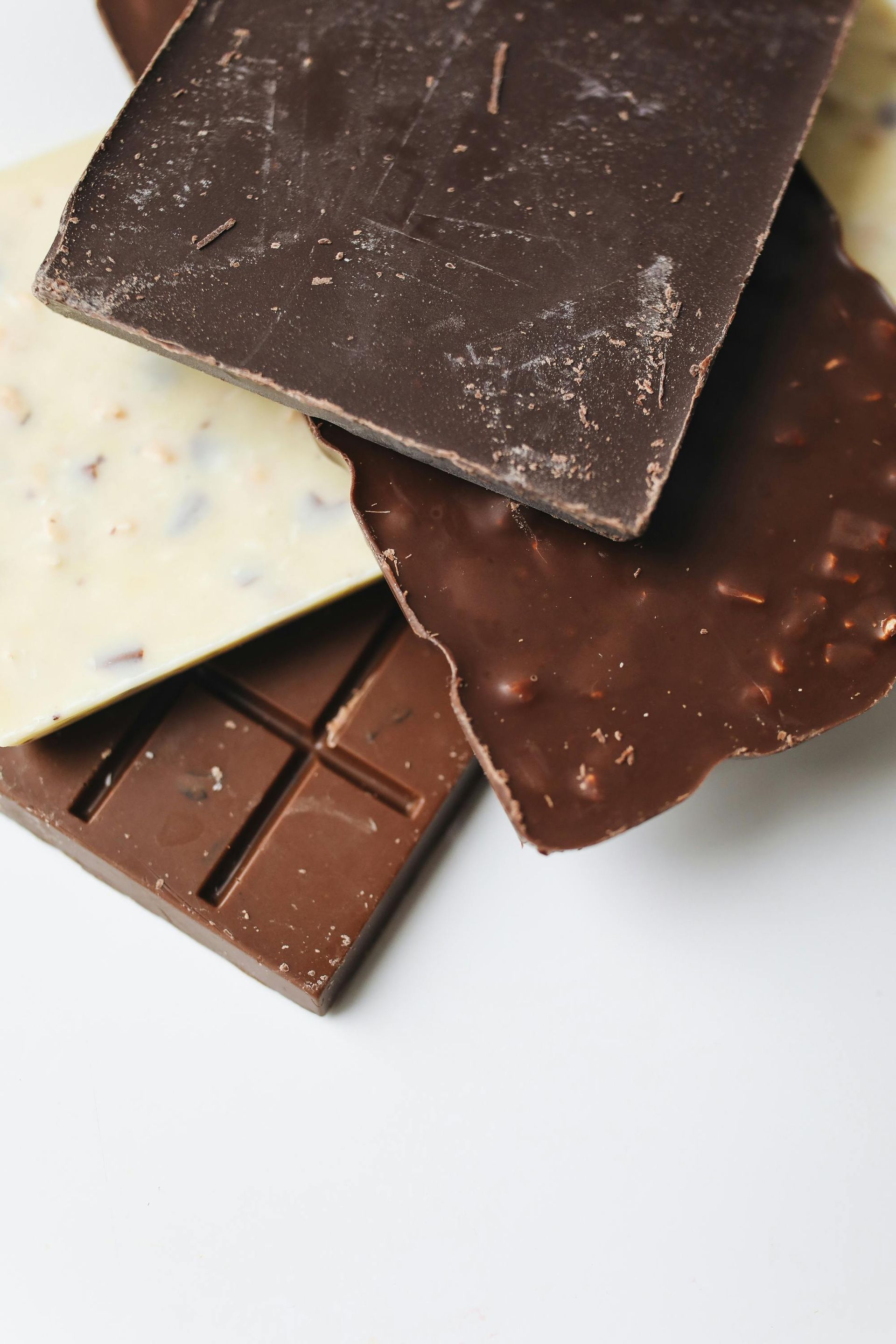 Vari pezzi di cioccolato, tra cui fondente, al latte e bianco, su una superficie bianca.