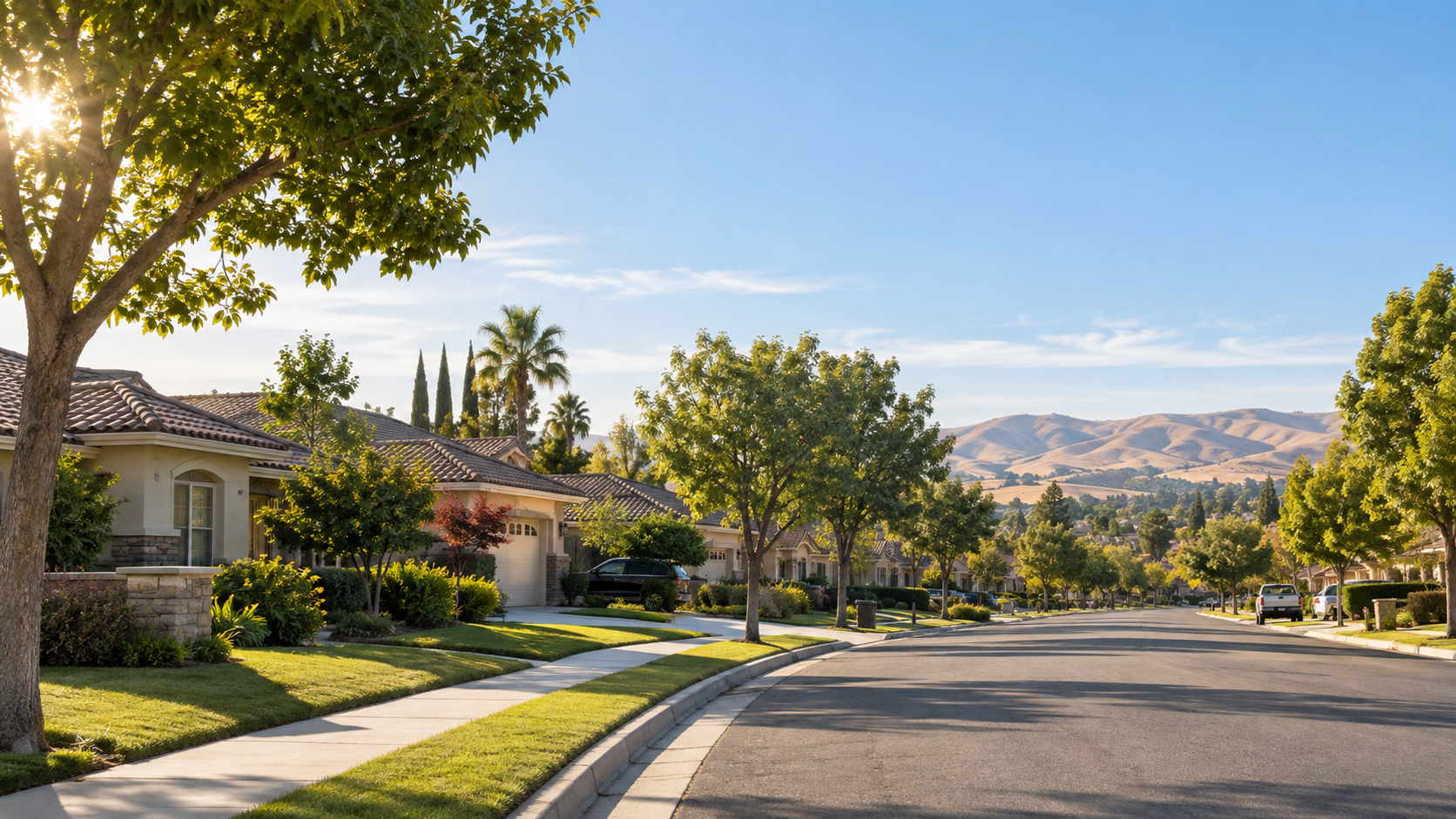 Quiet suburban street with trees, sidewalks, and houses under a clear blue sky with distant hills.