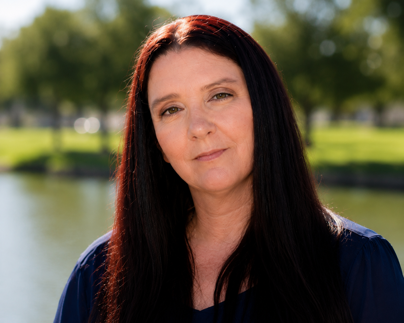 Portrait of a woman with long dark hair outdoors, with blurred trees and sunlight in the background