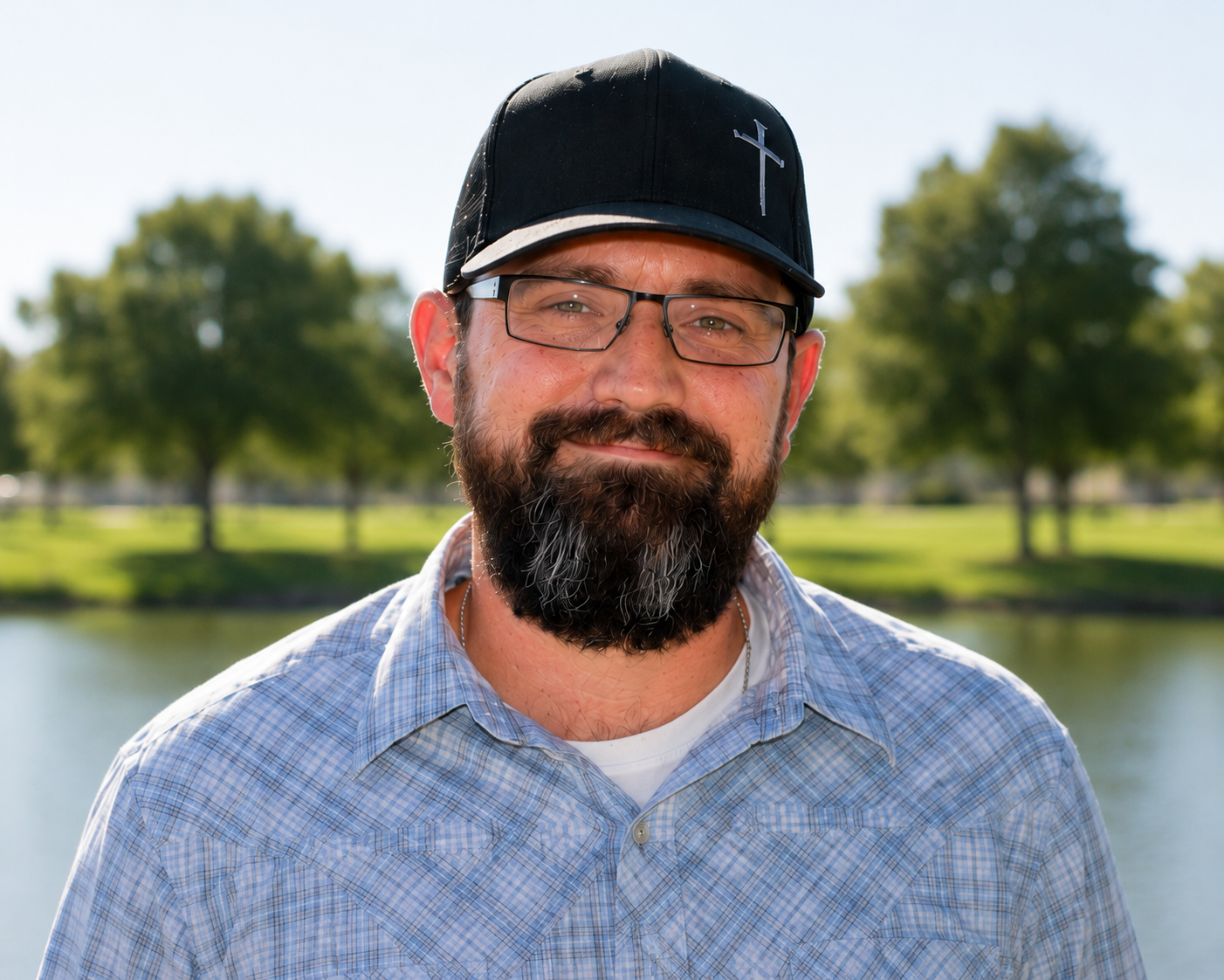 Smiling man in a black cap and glasses, standing outdoors in a sunlit park.