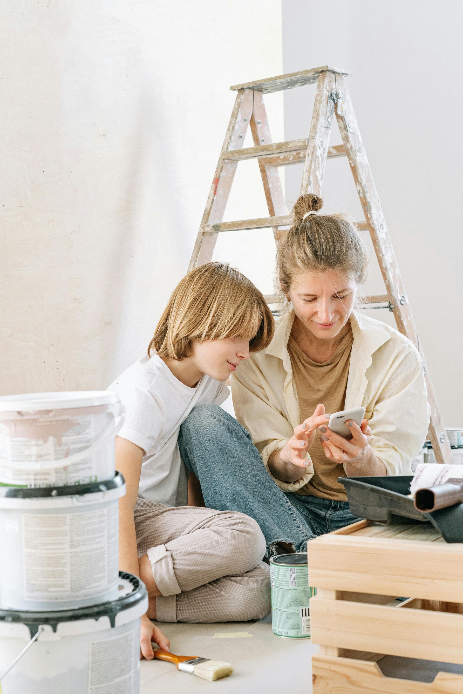 Woman and child looking at a phone while painting; buckets of paint and a ladder are visible.