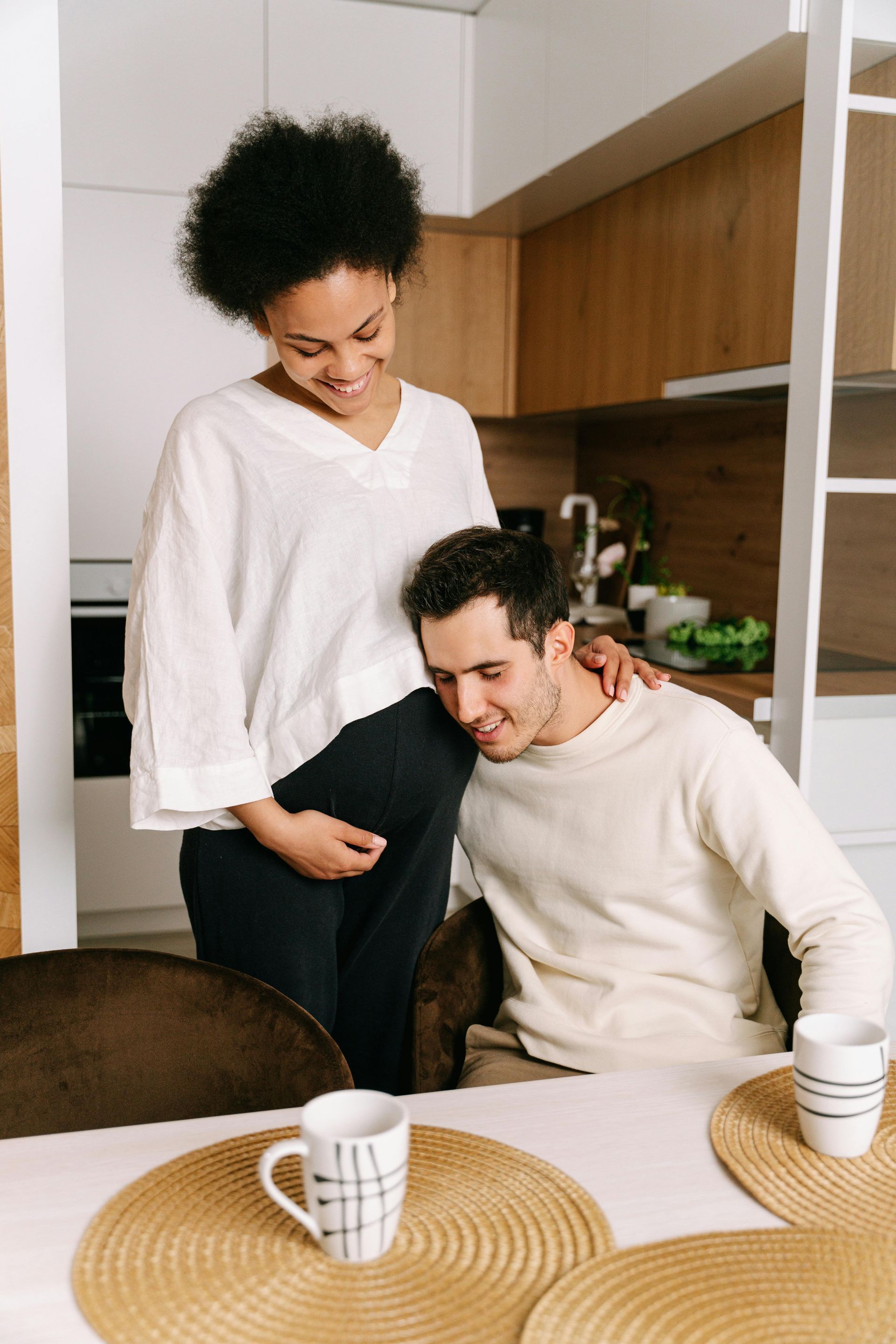 Pregnant woman smiles, holding her belly as man listens. They are in a kitchen, near a table.