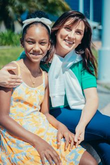 A young girl and woman smiling and embracing outdoors. The woman has her arm around the girl, who wears a yellow dress.