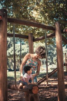 Woman helping a child on a wooden playground swing, in a sunny park setting.
