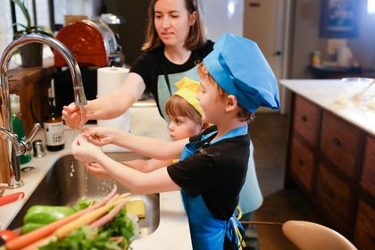 Woman and two children washing hands in a kitchen sink before cooking, wearing aprons and chef hats.