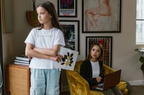 Girl with arms crossed, looks away from a woman working on a laptop in a yellow chair.