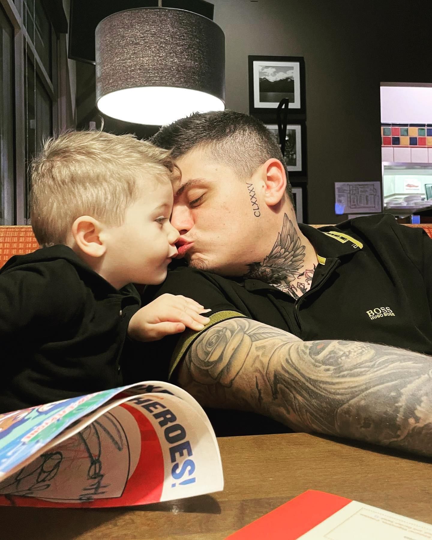 Man with tattoos kisses a young child on the cheek at a table in a restaurant.