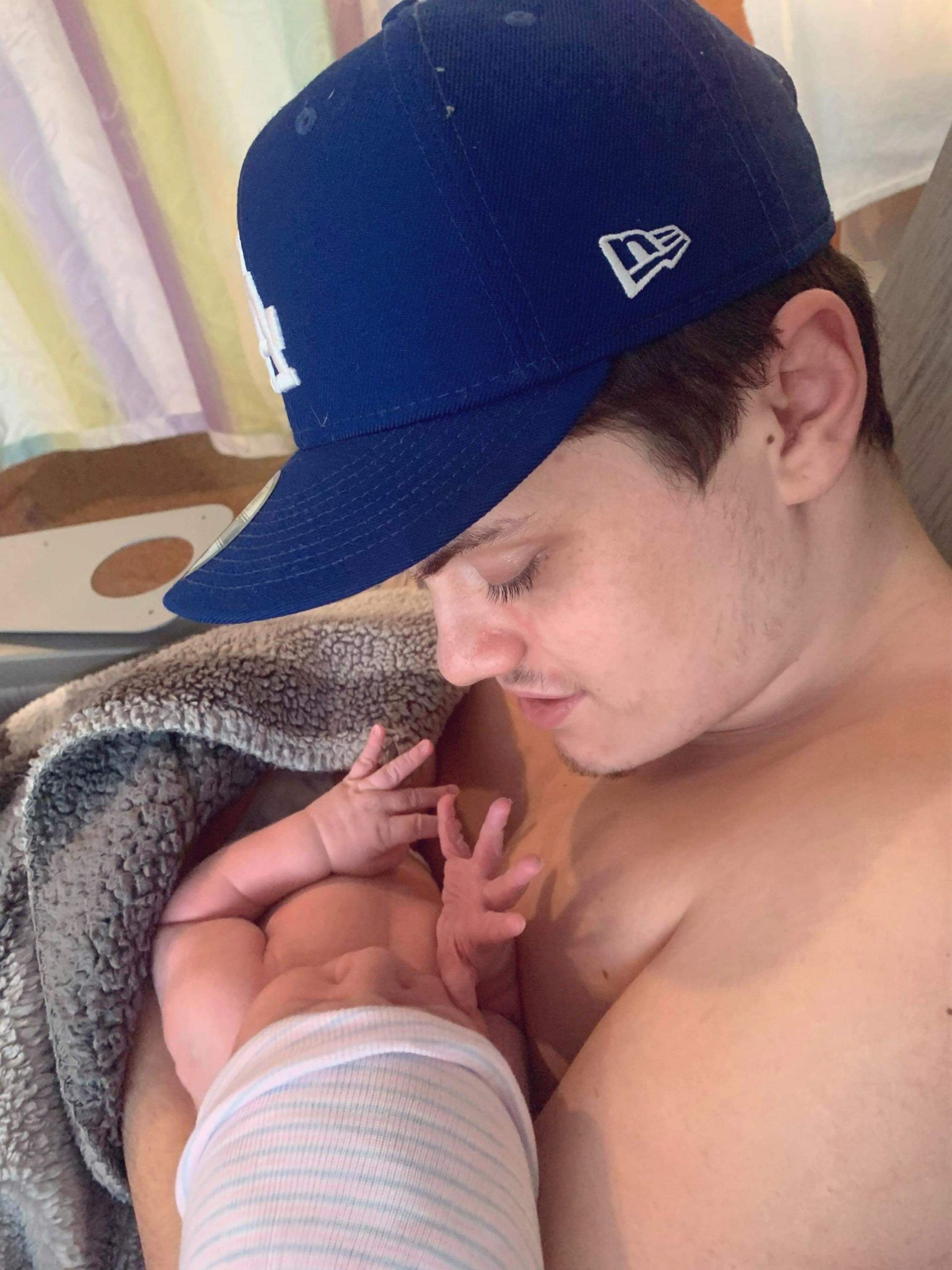 Man wearing blue cap, holding newborn baby. Baby's wrapped in white, man looks down, smiles softly.