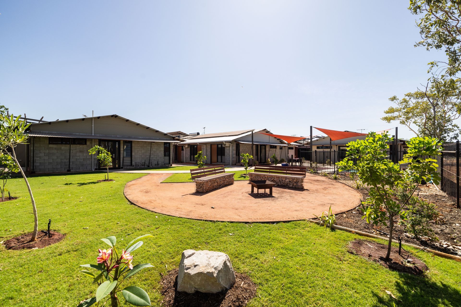 A sunny outdoor area with a circular gravel seating area, benches, and buildings in the background.