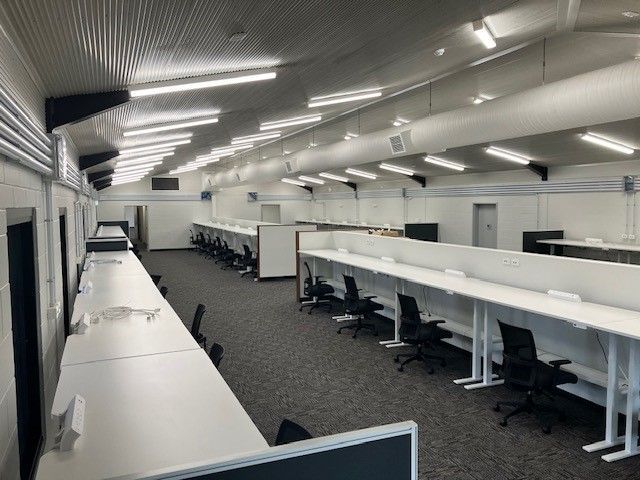 Empty call center with long white desks, black chairs, and grey carpet. Fluorescent lighting, white walls, and ductwork.
