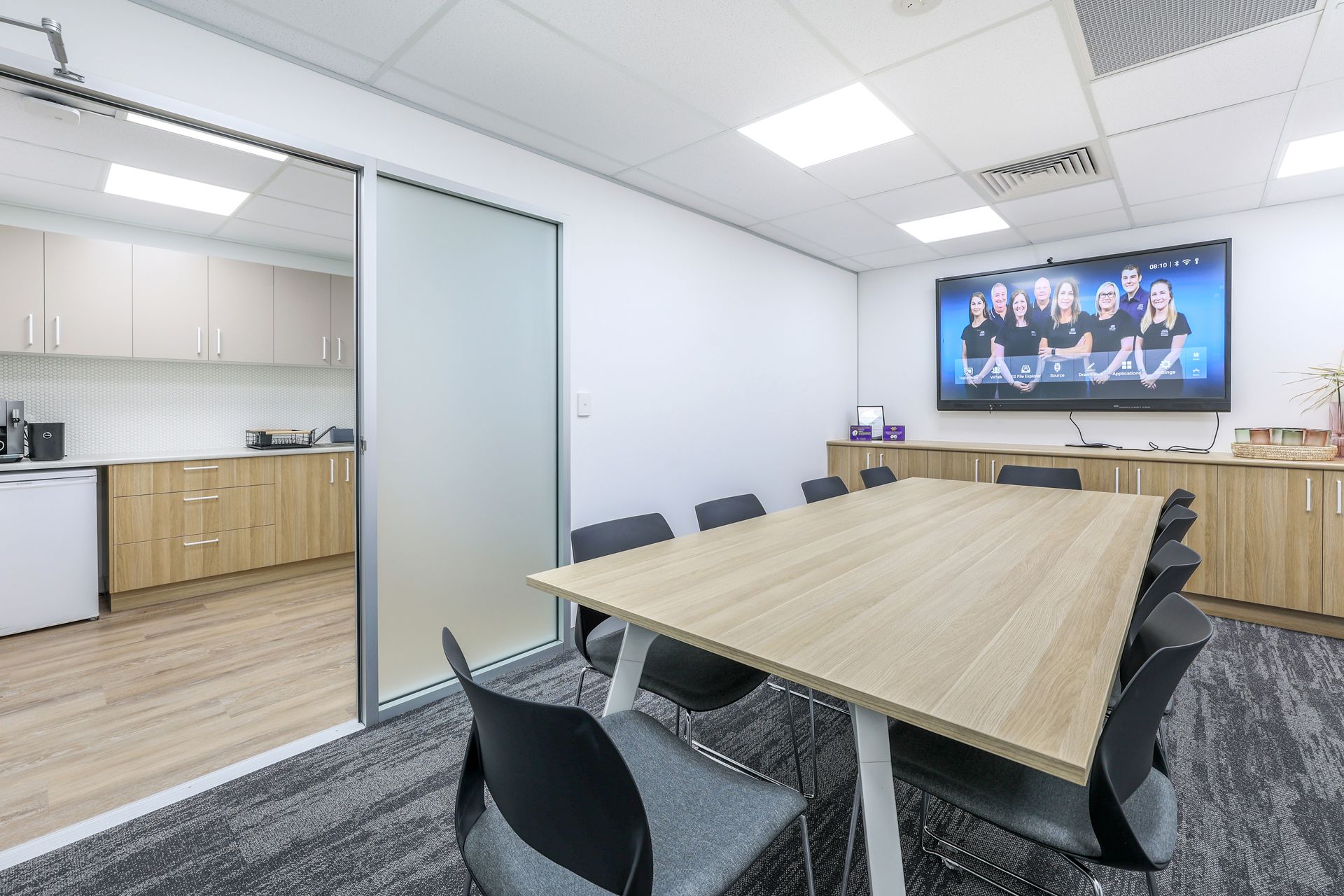Conference room with table, chairs, large screen TV, and kitchen area visible.